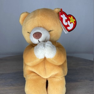 May include: A tan plush praying bear with a white belly and white paws. The bear is wearing a red Ty beanie baby tag with a white star and the words "Beanie Baby" on it.