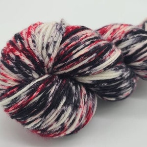 May include: A close-up of a skein of yarn with a speckled pattern. The yarn is a mix of red, black, white, and gray fibers. The yarn is tightly wound and appears soft, suitable for knitting or crochet projects.