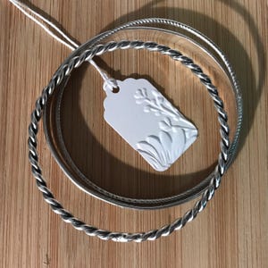 May include: A set of two silver-toned metal bangles, one featuring a twisted rope texture and the other a smooth, plain surface. A white rectangular paper tag with a scalloped top and an embossed floral design is attached by a white string.
