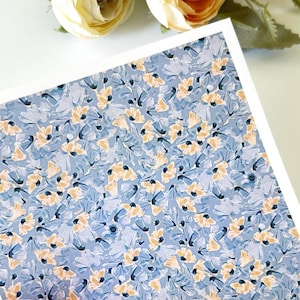 May include: A rectangular card with a blue floral pattern, held by a hand with a pink nail. The floral design features small yellow and blue flowers. Two cream-colored flowers and greenery are in the background.