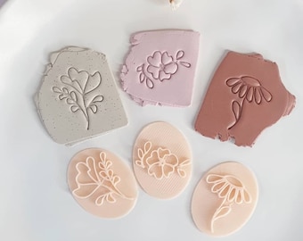 Puede incluir: Cinco sellos de arcilla polimérica con diseños florales. Dos son de forma ovalada y tres son de formas irregulares. Los sellos son en tonos de rosa, marrón y beige.
