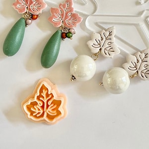 May include: Three pairs of leaf-shaped earrings with different colored beads and charms. The earrings are displayed on a white surface with a clear plastic hanger.
