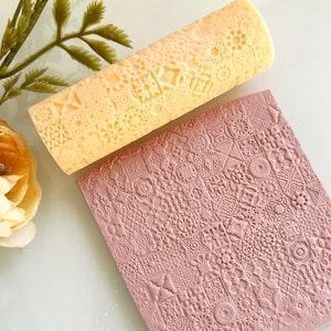 Op de afbeelding: Een lichtoranje deegroller en een rechthoekig roze kleiblad met een reliëf geometrisch patroon. De deegroller heeft een vergelijkbaar patroon. De items liggen op een wit oppervlak met een bloem en groen in de achtergrond.