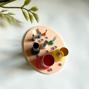May include: A set of metal and plastic circle cutters in various sizes, arranged on a light pink oval base. The cutters are in different colors, including red, yellow, and black. Numbers are printed on the base next to each cutter.