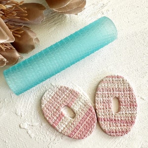 Puede incluir: Dos pendientes ovalados de color rosa y blanco con textura tejida y un agujero en el centro.