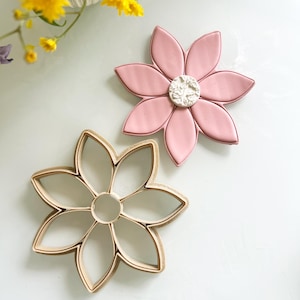 May include: Two flower-shaped cookie cutters. One is a light pink color with a white center, and the other is a light brown color. The cutters are on a white surface. Yellow flowers are in the background.