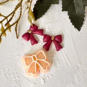 May include: Three red and gold bow-shaped polymer clay charms, one with a peach-colored outline.