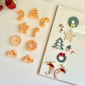 May include: Christmas-themed cookie cutters and earrings. The cutters are in shapes like trees, stars, and trucks. The earrings feature Santa Claus faces and crescent moons. The colour scheme includes peach, green, red, and white. Measurements are in centimetres.