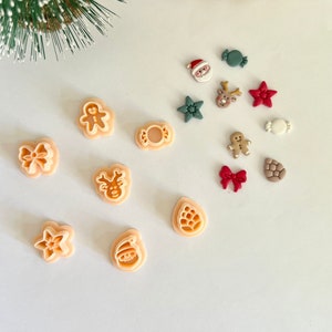 Peut inclure: Un ensemble de 12 emporte-pièces sur le thème de Noël de différentes formes, notamment une étoile, un renne, un bonhomme en pain d'épices, une canne en sucre, un nœud, un Père Noël, une cloche et un sapin. Les emporte-pièces sont en plastique orange clair et sont disposés sur une surface blanche.