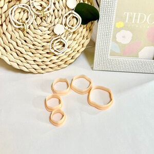 May include: A set of five white ceramic earrings with gold accents and a set of five peach-colored plastic cookie cutters in various sizes. The earrings and cookie cutters are displayed on a white surface with a woven basket and a white picture frame in the background.