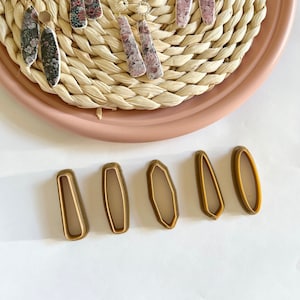 Op de afbeelding: Een set van 10 oorringen met bloemenpatronen en gouden accenten, gepresenteerd op een geweven rieten bord. De oorringen zijn in paren gerangschikt, waarbij elk paar een ander bloemenpatroon heeft. Het bord staat op een roze oppervlak. Er zijn ook vier goudkleurige koekjesuitstekers in de vorm van een ovaal op een wit oppervlak.