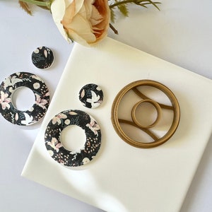 Pode incluir: Três brincos redondos com padrão floral e um cortador de anéis de metal dourado. Os brincos são pretos com flores rosa e brancas.