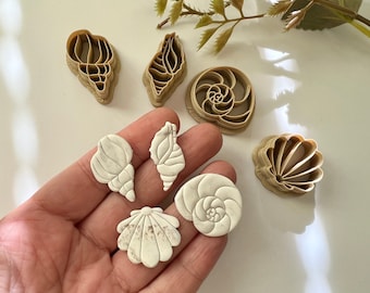 Peut inclure: Un ensemble de six emporte-pièces en plastique brun en forme de coquillages. Les emporte-pièces sont sur une surface blanche et une main tient cinq coquillages en argile blanche qui ont été découpés avec les emporte-pièces.