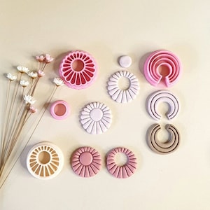 Op de afbeelding: Een set roze en witte polymer clay oorbellen met een madeliefje-ontwerp, samen met de mallen die gebruikt werden om ze te maken. De mallen zijn gemaakt van wit plastic en hebben een madeliefje-ontwerp.