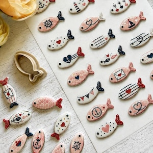 May include: A collection of small, flat, fish-shaped ceramic or clay pieces featuring various nautical and heart-themed patterns in pink, white, navy blue, and red. The set includes a single fish-shaped cookie cutter and loose pieces.