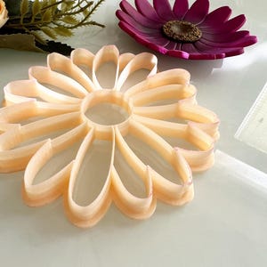 May include: A light orange flower-shaped cookie cutter with a central hole, next to a purple flower-shaped dish with a brown centre. Artificial flowers are in the background.