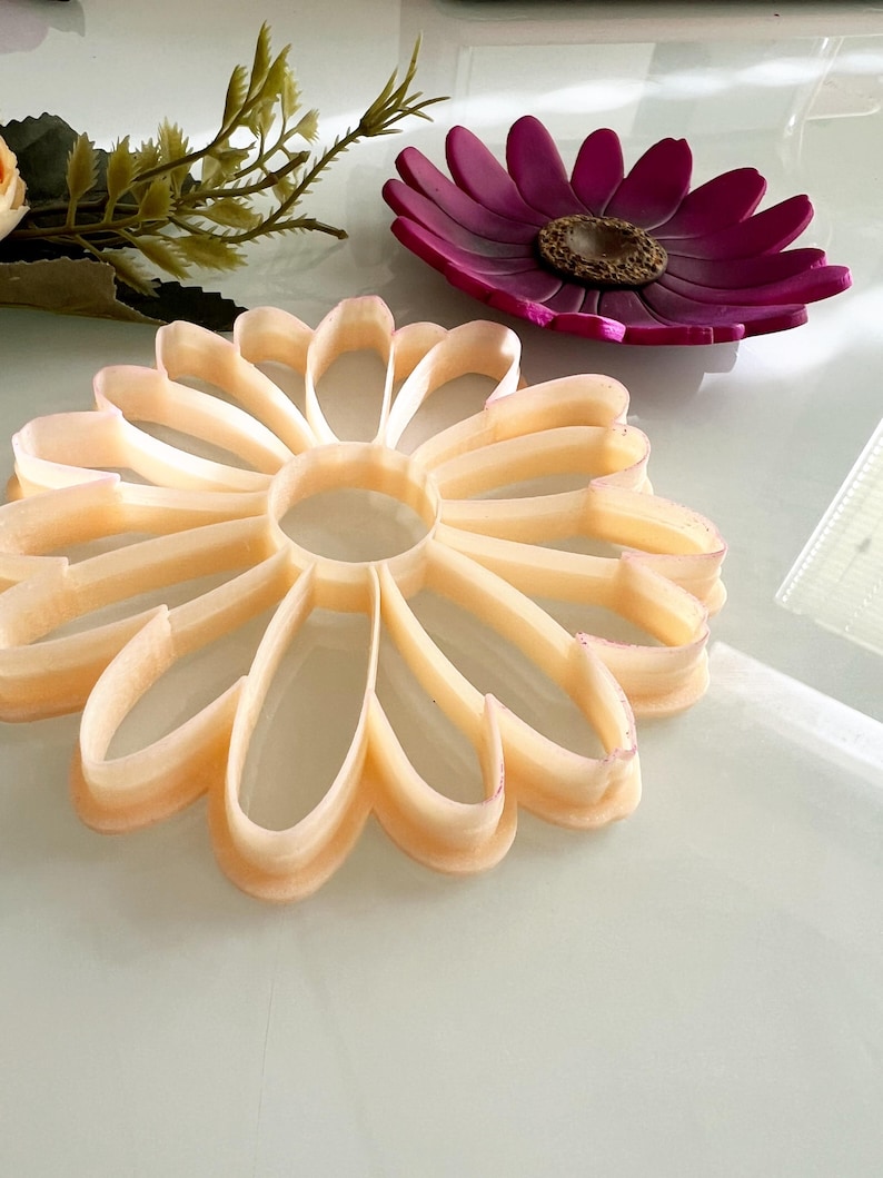 May include: A light orange flower-shaped cookie cutter with a central hole, next to a purple flower-shaped dish with a brown centre. Artificial flowers are in the background.