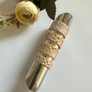 Pode incluir: Um tampão de garrafa de vinho em aço inoxidável com cinco faixas bege texturizadas. As faixas têm um padrão pontilhado. O tampão é cilíndrico e tem uma extremidade curva. Flores decorativas no fundo.