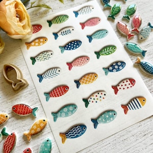 May include: A collection of small, flat, fish-shaped ceramic pieces featuring a variety of colorful patterns including stripes, polka dots, floral motifs, and geometric designs. The fish are arranged on a white rectangular sheet, with additional loose pieces scattered around, alongside a single beige plastic fish-shaped cookie cutter.