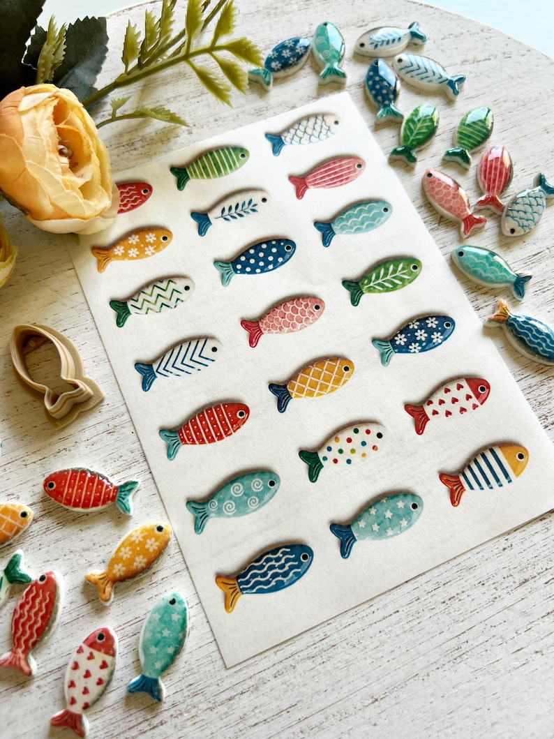 May include: A collection of small, flat, fish-shaped ceramic pieces featuring a variety of colorful patterns including stripes, polka dots, floral motifs, and geometric designs. The fish are arranged on a white rectangular sheet, with additional loose pieces scattered around, alongside a single beige plastic fish-shaped cookie cutter.