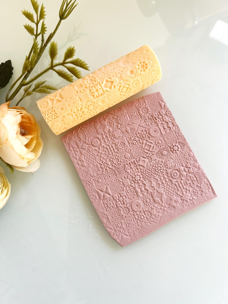 Pu&ograve; includere: Un mattarello arancione chiaro e un foglio di argilla rosa rettangolare con un motivo geometrico in rilievo. Il mattarello ha un motivo simile. Gli oggetti sono su una superficie bianca con un fiore e del verde sullo sfondo.