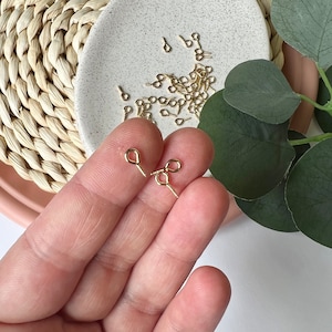 Può includere: Spille per occhi dorate con un anello in cima e un'estremità appuntita. Le spille per occhi sono sparse su una superficie bianca e una mano ne tiene due.