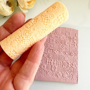 Pu&ograve; includere: Un mattarello beige chiaro con un motivo dettagliato e ripetuto &egrave; tenuto sopra una superficie rettangolare malva con un design in rilievo abbinato. Il mattarello &egrave; cilindrico e presenta una variet&agrave; di piccole forme geometriche.