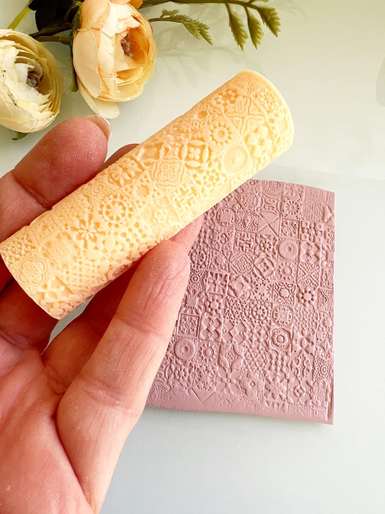 Pu&ograve; includere: Un mattarello beige chiaro con un motivo dettagliato e ripetuto &egrave; tenuto sopra una superficie rettangolare malva con un design in rilievo abbinato. Il mattarello &egrave; cilindrico e presenta una variet&agrave; di piccole forme geometriche.