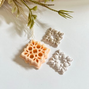 May include: Three decorative, square-shaped, floral embellishments. One is a light peach color with a detailed, intricate design. Another is a silver color with a simple floral design. The third is white with a simple floral design.