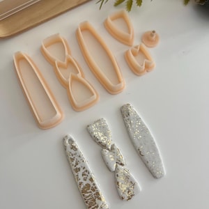 Könnte beinhalten: Eine Sammlung von Ohrringkomponenten und Ausstechern aus Polymer Clay. Die Ausstecher sind pfirsichfarben und haben verschiedene Formen. Die fertigen Ohrringe sind weiß mit Goldblatt-Akzenten. Die Komponenten umfassen lange, ovale und Schleifenformen.