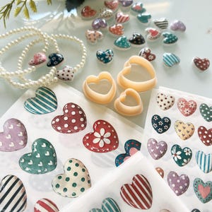 May include: A collection of heart-shaped items including small ceramic heart beads with various patterns, three beige plastic heart-shaped cutters, and sheets of heart-themed stickers featuring stripes, polka dots, and floral designs.