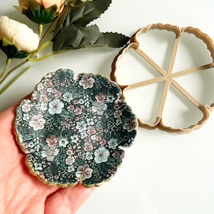 May include: A floral-patterned coaster with a dark background and gold trim, next to a matching cookie cutter. The coaster features a delicate pattern of pink, white, and blue flowers. Artificial peach-colored roses and greenery are in the background.