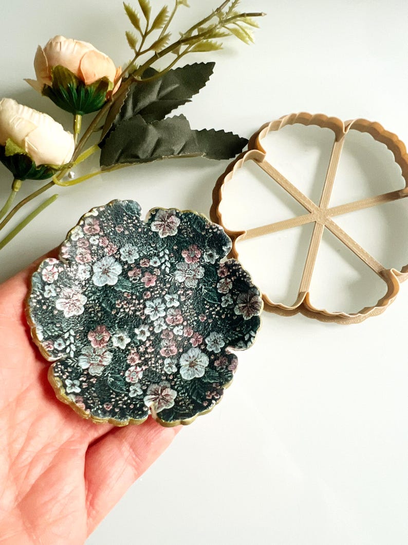 May include: A floral-patterned coaster with a dark background and gold trim, next to a matching cookie cutter. The coaster features a delicate pattern of pink, white, and blue flowers. Artificial peach-colored roses and greenery are in the background.