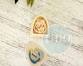 May include: A beige cookie cutter in the shape of a nativity scene, next to a painted ceramic piece with a similar design. The ceramic piece features blue, yellow, and white colors. The background is a white wooden surface with green leaves.