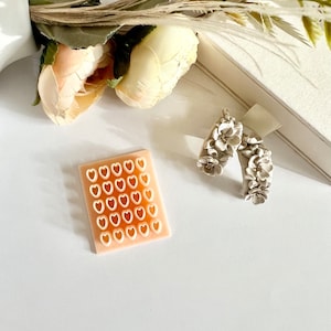 May include: A sheet of clear heart-shaped silicone moulds on a peach-coloured background. The moulds are arranged in a grid pattern. There are two white floral earrings in the background.