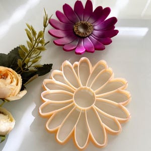 May include: Two daisy-shaped objects are shown. The top one is a deep magenta with a brown centre. The bottom one is a light peach colour. They appear to be cookie cutters or craft supplies.