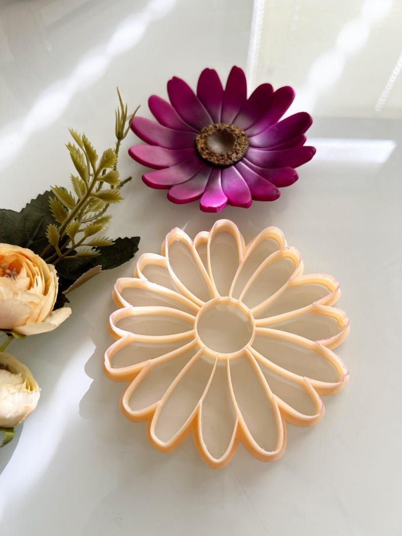 May include: Two daisy-shaped objects are shown. The top one is a deep magenta with a brown centre. The bottom one is a light peach colour. They appear to be cookie cutters or craft supplies.