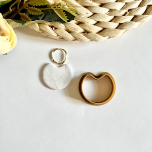 Op de afbeelding: Een goudkleurige ring, een witte steen met marmerpatroon en een goudkleurig hartvormig object zijn gerangschikt op een wit oppervlak. Dit zijn waarschijnlijk sieraden of decoratieve items. Een gevlochten mand en groen zijn op de achtergrond.