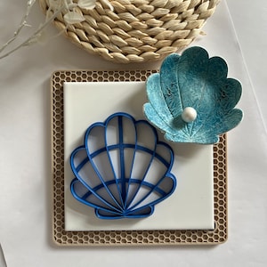 May include: A blue plastic seashell cookie cutter with a white pearl on a blue and white seashell dish. The seashells are on a beige silicone baking mat with a honeycomb pattern.