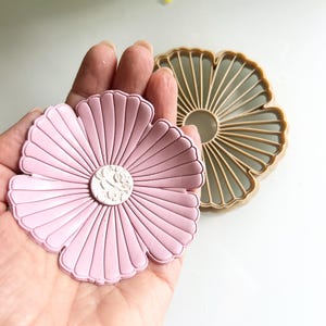 May include: A pink flower-shaped cookie cutter with a detailed center design is held in a hand. Another tan-colored flower-shaped cutter is next to it. The cutters are on a white surface, with yellow flowers in the background.