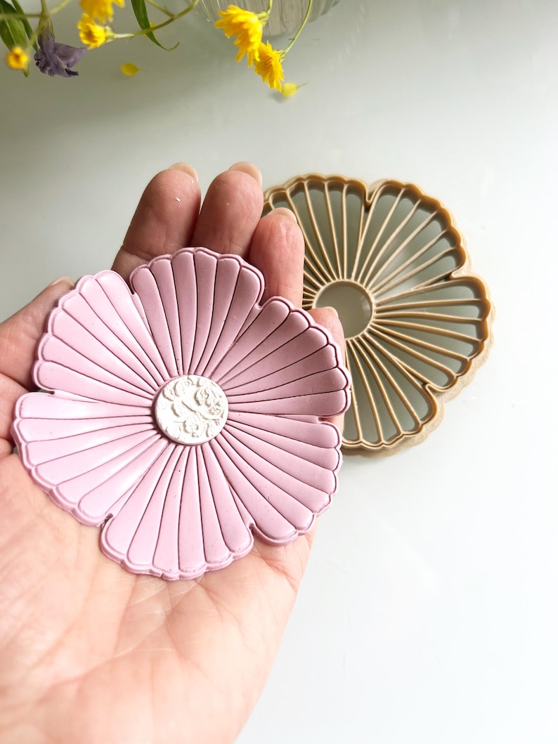 May include: A pink flower-shaped cookie cutter with a detailed center design is held in a hand. Another tan-colored flower-shaped cutter is next to it. The cutters are on a white surface, with yellow flowers in the background.
