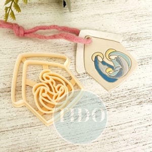 Puede incluir: Una etiqueta de cerámica beige con una escena de la Natividad en azul y dorado, colgada de un cordón rosa, junto a un cortador de galletas beige con forma de rosa. La etiqueta y el cortador están sobre una superficie de madera blanca envejecida.