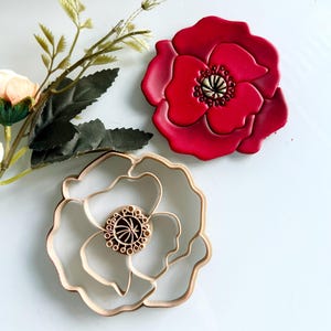 May include: A vibrant red poppy-shaped cookie cutter and a red poppy-shaped cookie. The cookie cutter is made of light-colored wood. The poppy cookie has a detailed center and is next to a sprig of greenery.
