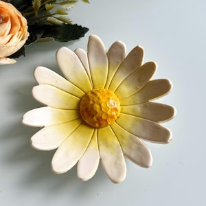 May include: A ceramic daisy-shaped dish with white petals and a yellow centre. The petals have a slight yellow gradient. A peach-coloured flower and green leaves are visible in the background. This decorative dish is ideal for holding small items.