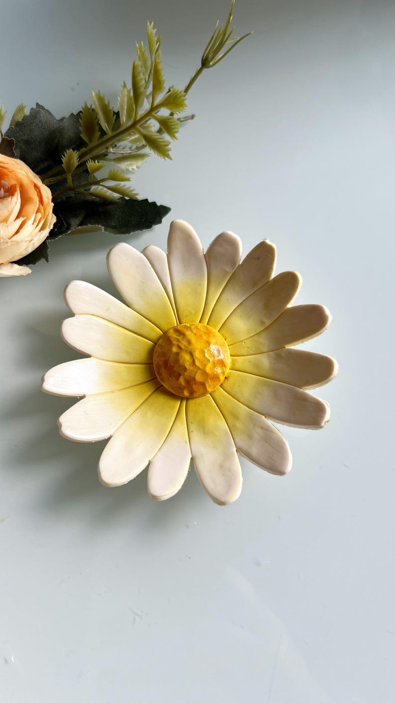 May include: A ceramic daisy-shaped dish with white petals and a yellow centre. The petals have a slight yellow gradient. A peach-coloured flower and green leaves are visible in the background. This decorative dish is ideal for holding small items.