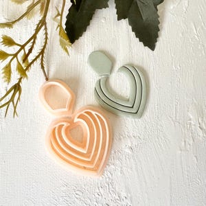 Peut inclure: Deux paires de boucles d'oreilles en forme de cœur et un emporte-pièce en forme de cœur. Une paire de boucles d'oreilles est vert clair avec des rayures noires, l'autre est de couleur pêche. L'emporte-pièce est de couleur pêche avec des couches en forme de cœur.