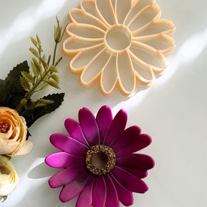 May include: Two decorative flower-shaped items. One is a light orange colour with a petal design. The other is a deep magenta with a textured, brown centre. The flowers are on a white surface.