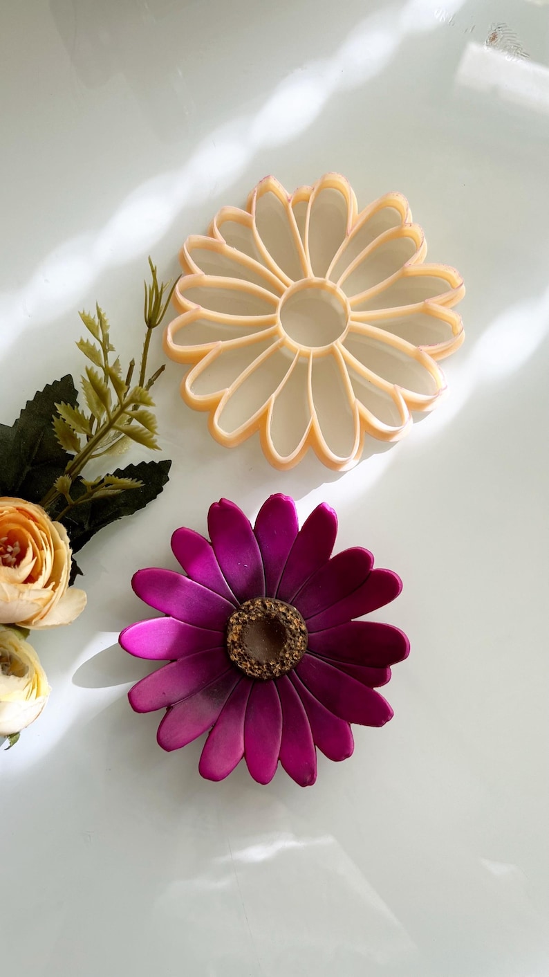 May include: Two decorative flower-shaped items. One is a light orange colour with a petal design. The other is a deep magenta with a textured, brown centre. The flowers are on a white surface.