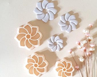 May include: Set of six cookie cutters in the shape of a flower with a swirl design. The cutters are in two colors, light gray and light peach.