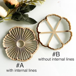 May include: Two wooden flower-shaped cookie cutters. One has internal lines, labeled "#A with internal lines." The other, labeled "#B without internal lines," lacks internal lines. Artificial flowers and leaves are in the background.